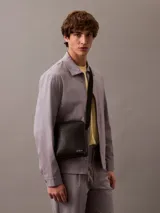 Bandolera Calvin Klein Jeans negra de poliuretano con correa ajustable, cierre superior con cremallera y logo estampado en el frente.