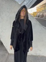 Bata de diseño holgado confeccionada en tela de textura suave y aterciopelada, color negro. Presenta una capucha amplia que puede utilizarse como bufanda, puños elásticos ajustados y bolsillos laterales.