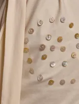 Vestido corto de color beige con diseño asimétrico de un solo hombro y manga corta, adornado con patrones circulares de botones decorativos. Incluye un lazo ajustable para la cintura.