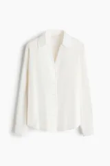 Blusa blanca de tela con brillo, cuello clásico, escote en V con botones frontales, mangas largas con puños sencillos y bajo redondeado.