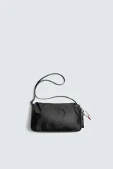 Bolso tipo bandolera de diseño minimalista en color negro, con cierre de cremallera superior y asa de hombro ajustable. Incluye un detalle decorativo de charms metálicos en uno de los laterales.