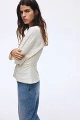 Top de manga larga confeccionado en punto de algodón, con cuello redondo, hombros caídos y detalle de frunces laterales que crean un efecto drapeado, finalizando en un bajo acampanado.