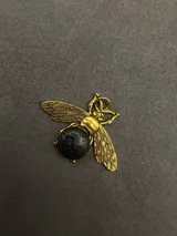 Dije con forma de abeja en metal dorado, con alas detalladas y cuerpo decorado con pequeñas piedras naturales facetadas en tonos variados.