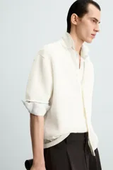 Camisa de hombre de corte relajado, color blanco roto, confeccionada en tejido de punto con estructura abierta y textura acanalada. Presenta cuello solapa y manga corta.