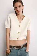 Chaqueta corta de punto texturizado color blanco roto, con escote en V, manga corta y cierre frontal con tres botones metálicos dorados en forma de estrella de mar.