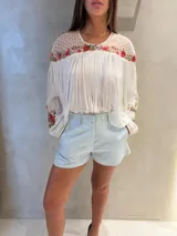 Blusa blanca de manga larga con detalles semitransparentes de lunares, bordados florales multicolores en el pecho y las mangas, y flecos largos colgantes en el frente.