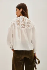 Camisa blanca de estilo romántico con cuello alto, confeccionada en algodón con detalles de encaje y bordados en el frente. Presenta mangas largas abullonadas con puños elásticos y terminaciones de encaje.