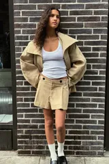 Trench color beige con cuello de solapa subido y manga larga. Tiene bolsillos laterales, forro interior y cinturón de lazo en el mismo tejido. El cuello se cierra con un botón.