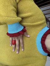 Buzo de punto oversize color rojo, con cuello alto y puños con terminaciones en volados. Presenta detalles en contraste color amarillo en el cuello y los puños, con un tejido de acabado ligeramente brillante.