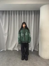 Buzo con capucha y cierre frontal completo, color verde oscuro con efecto lavado degradado. Presenta estampado gráfico en tipografía estilizada en el frente y mangas, bolsillos laterales y etiqueta de marca en el puño.