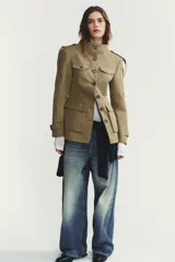 Chaqueta de estilo militar en color beige, con cuello subido, cierre frontal de botones, bolsillos de plastrón con solapa en el delantero y detalle de solapas en el pecho. Presenta mangas largas con hombreras y trabillas decorativas.