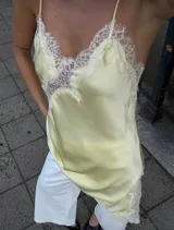 Vestido corto de satén color amarillo pastel con diseño asimétrico. Presenta detalles de encaje blanco en el escote, lateral y ruedo, con breteles finos ajustables.