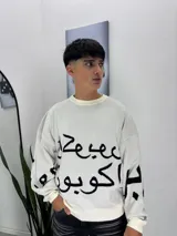 Sweater de punto color beige con estampado de caligrafía árabe en color negro. Presenta cuello redondo, puños y ruedo acanalados, con un diseño de corte holgado.