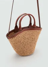 Bolso tipo cesta de fibras naturales con diseño en forma de abanico. Presenta detalles en cuero sintético marrón en el borde superior, asas cortas de mano y una correa bandolera larga ajustable. Incluye cierre de botón magnético y forro interior.
