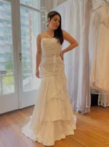 Vestido de novia blanco, largo hasta el piso, con breteles finos y escote recto. Presenta un fajín con apliques de strass en la cintura y falda con volados.