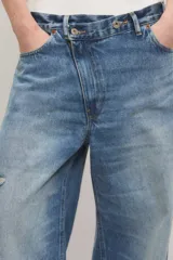 Jeans de denim azul con calce holgado y tiro medio, con diseño de cintura asimétrica cruzada, roturas localizadas y ruedo desflecado con degradé.