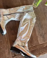 Bota alta plateada de cuero sintético con suela track negra, cierre lateral con cremallera y detalle de hebilla ajustable en el tobillo.