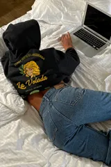 Canguro negro de corte oversize con capucha y bolsillo frontal. Presenta un diseño gráfico estampado en la espalda que simula un paquete de yerba mate, con la ilustración de una mujer, hojas y la inscripción 'La Volada'.