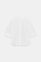 Camisa blanca de algodón con cuello redondo y manga corta abullonada. Presenta un canesú con pliegues verticales en la parte delantera y cierre frontal con botones ocultos. El bajo es redondeado.