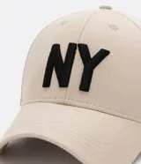 Gorra de seis paneles con ala curva, confeccionada en algodón color marrón. Presenta un bordado frontal con las letras NY en color blanco y cuenta con cierre strapback ajustable en la parte posterior.