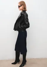 Chaqueta tipo biker de poliuretano color negro, con solapas amplias, cierre frontal cruzado con cremallera, bolsillos laterales con cierre y trabillas en los hombros.