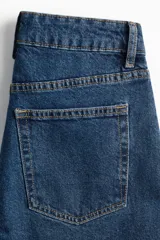 Jean tobillero de denim azul medio, con cinco bolsillos, tiro alto y cierre de botón. Presenta un corte slim fit ajustado en cadera y muslo, con pierna recta.