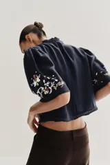 Top corto de color negro con escote en pico y mangas cortas anchas. Presenta bordados coloridos en estilo geométrico y floral, detalle de entredoses y cierre frontal mediante lazos con abalorios.