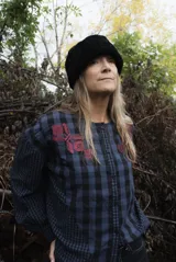 Blusa de algodón con estampado a cuadros en tonos azul y negro, con bordados decorativos en color bordó sobre el canesú. Presenta cierre frontal con botones, cuello redondo y mangas largas levemente abullonadas.