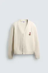 Cárdigan de punto color beige con cuello en pico, manga larga y cierre frontal de botones. Presenta un pequeño bordado a contraste en el pecho y acabados en rib en puños y bajo.