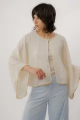 Blusa de lino color beige, de corte amplio y mangas largas acampanadas, con detalles de bordado en los bordes y cierre frontal superior con un botón.