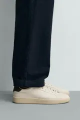 Zapatilla deportiva de diseño minimalista en color crema, con detalle de contraste en color marrón oscuro en el talón y suela de goma con volumen.
