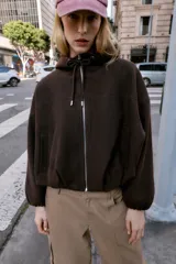 Chaqueta estilo bomber de corte oversize en color marrón oscuro, confeccionada en tejido suave. Presenta capucha con cordones ajustables, cierre frontal con cremallera, mangas largas con puños elásticos y bolsillos delanteros de vivo. El bajo cuenta con detalle de pliegues.