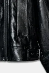 Chaqueta de estilo biker confeccionada en tejido efecto piel con acabado brillante. Presenta cuello con solapas, cierre frontal con cremallera y botones, bolsillos delanteros y bajo ajustable con trabillas y hebilla metálica.