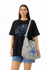 Bolso de tela tipo tote bag, con estampado de rayas verticales en tonos beige y negro/gris. Presenta un estampado frontal en azul que incluye una figura ecuestre bajo una palmera.