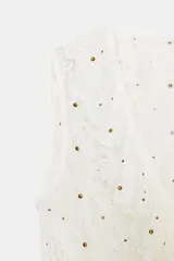 Top sin mangas color blanco roto, con escote en pico y detalle de volantes en los bordes. Presenta pequeñas tachas doradas distribuidas por toda la prenda y cierre frontal con lazadas. El bajo es asimétrico.