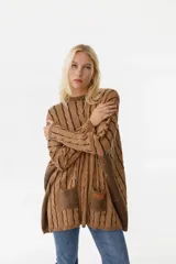 Sweater de punto color camel con diseño de trenzas verticales y acabado brillante. Presenta una silueta holgada tipo poncho, cuello redondo, mangas largas con puños ajustados y bolsillos frontales con detalle de etiqueta de cuero.