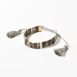 Pulsera tejida con cuentas rectangulares en tonos gris, marrón y blanco, con detalles dorados. Ajustable con cordón gris y terminaciones con borlas.
