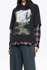 Camiseta negra de corte oversize con cuello redondo y mangas cortas. Presenta un estampado gráfico grande en el frente que simula un paisaje nocturno o crepuscular con un ciervo, árboles y montañas.