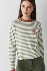 Remera de manga larga con diseño a rayas horizontales en color verde y blanco, cuello redondo en color marrón y pequeño bordado circular en el pecho.