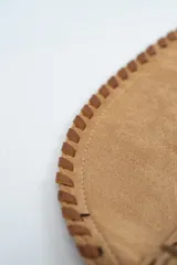 Bolso tipo hobo de gamuza color camel, con flecos largos laterales, correa de hombro de cuero sintético y cierre central con solapa y botón a presión.