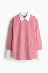 Camisa oversized de popelina de algodón con diseño a rayas verticales rojas y blancas. Presenta cuello clásico y puños anchos en color blanco sólido, cierre frontal con botones y bajo redondeado con aberturas laterales.