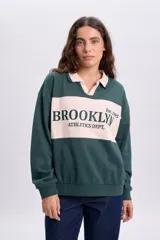 Sudadera polo verde oscuro con cuello y franja central en color beige. Presenta un diseño de manga larga y cuenta con un bordado frontal con el texto "BROOKLYN ATHLETICS DEPT. Est. 1995".
