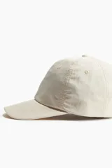 Gorra color crema de sarga de algodón con ojales bordados y correa ajustable con hebilla metálica en la parte posterior.
