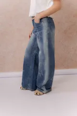 Jean de tiro medio y pierna amplia, con calce wide leg y lavado azul con efecto desgastado. Presenta estampado floral y de mariposas en toda la prenda, bolsillos delanteros y traseros, y cierre frontal.