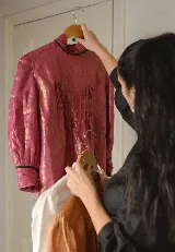 Blusa de lino con cuello alto, alforzas frontales y mangas abullonadas con puños terminados en puntilla negra. Presenta cierre posterior con botones a la vista.