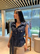 Chaleco de denim azul oscuro, sin mangas, con cuello camisero, cierre frontal de botones dorados y cinturón de lazo a juego.