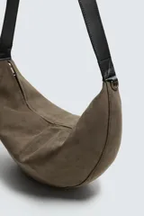 Bolso de hombro con diseño de media luna, confeccionado en gamuza color marrón. Cuenta con un compartimento principal con cierre de cremallera y un bolsillo interior con cierre. Posee un asa de hombro integrada.