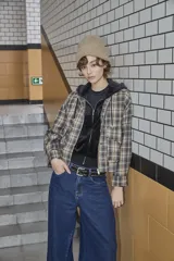 Chaqueta de estilo camisa a cuadros en tonos beige y marrón, con cierre frontal, cuello camisero y capucha integrada. Presenta un diseño holgado y mangas largas.