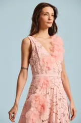 Vestido largo de encaje color rosado, con escote en V y aplicaciones de flores de tela tridimensionales en cascada sobre el hombro y el lateral de la falda. Incluye un cinturón delgado integrado en la misma tela.