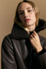 Campera tipo gamulán de cuero sintético negro, con interior y detalles de corderito sintético al tono en cuello y ruedo. Presenta cierre frontal metálico y diseño de silueta relajada.